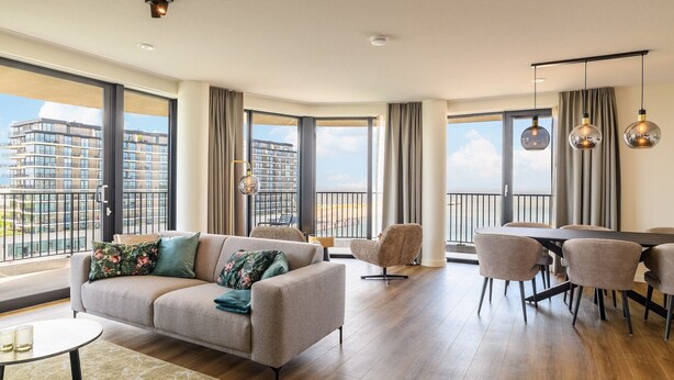 Un salon spacieux comprenant un canapé, des fauteuils et un coin repas. De grandes fenêtres offrent une vue sur les bâtiments et donnent accès à un balcon.