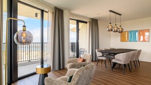 Un salon moderne doté d’une grande porte-fenêtre coulissante donnant sur un balcon, d’un coin repas avec une table noire et des chaises grises, et d’un lustre.