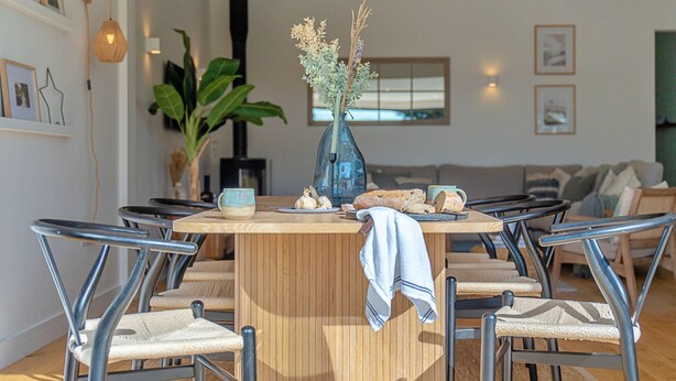 A dining area with a wooden table, six chairs, a vase with dried plants, and a window in the background. - Firethorn (Bude)