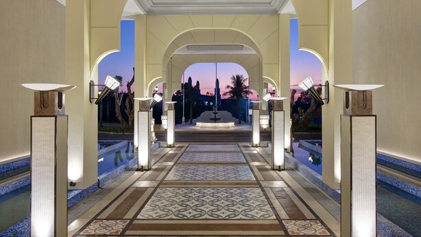 A grand hallway with a tiled floor, leading to a fountain and a view of a city skyline at dusk.