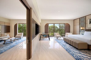 A modern hotel room with a large bed, a seating area with sofas and chairs, a dining table, and large windows offering a view of the outdoors.