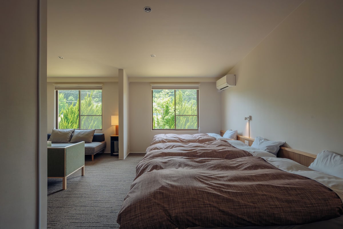 Een slaapkamer met een groot bed, twee ramen, een nachtkastje, een lamp en een zithoek met een bank en een kleine tafel.