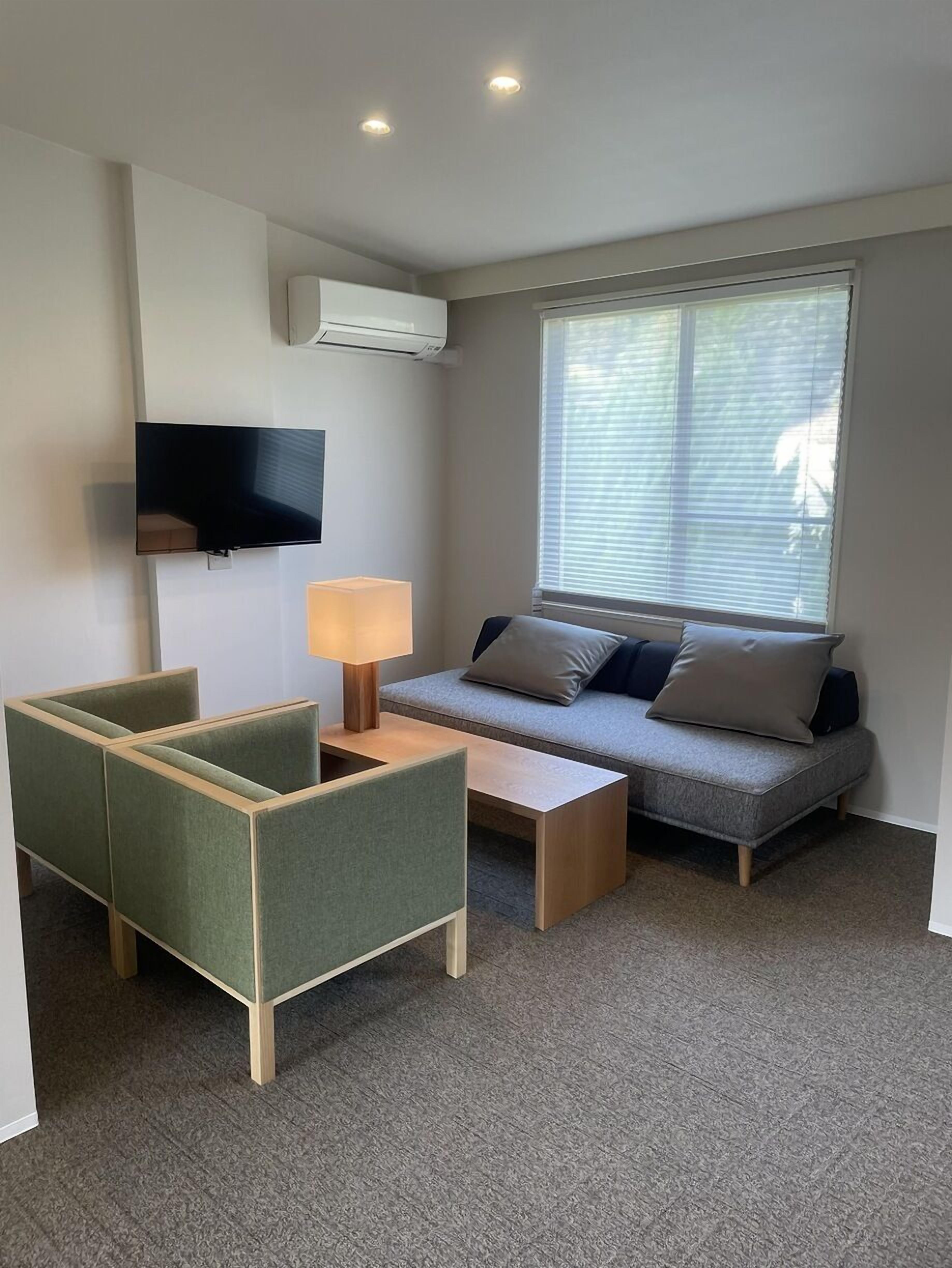 Un salon moderne comprenant un canapé, deux fauteuils, une table basse, un téléviseur et une fenêtre avec des stores.