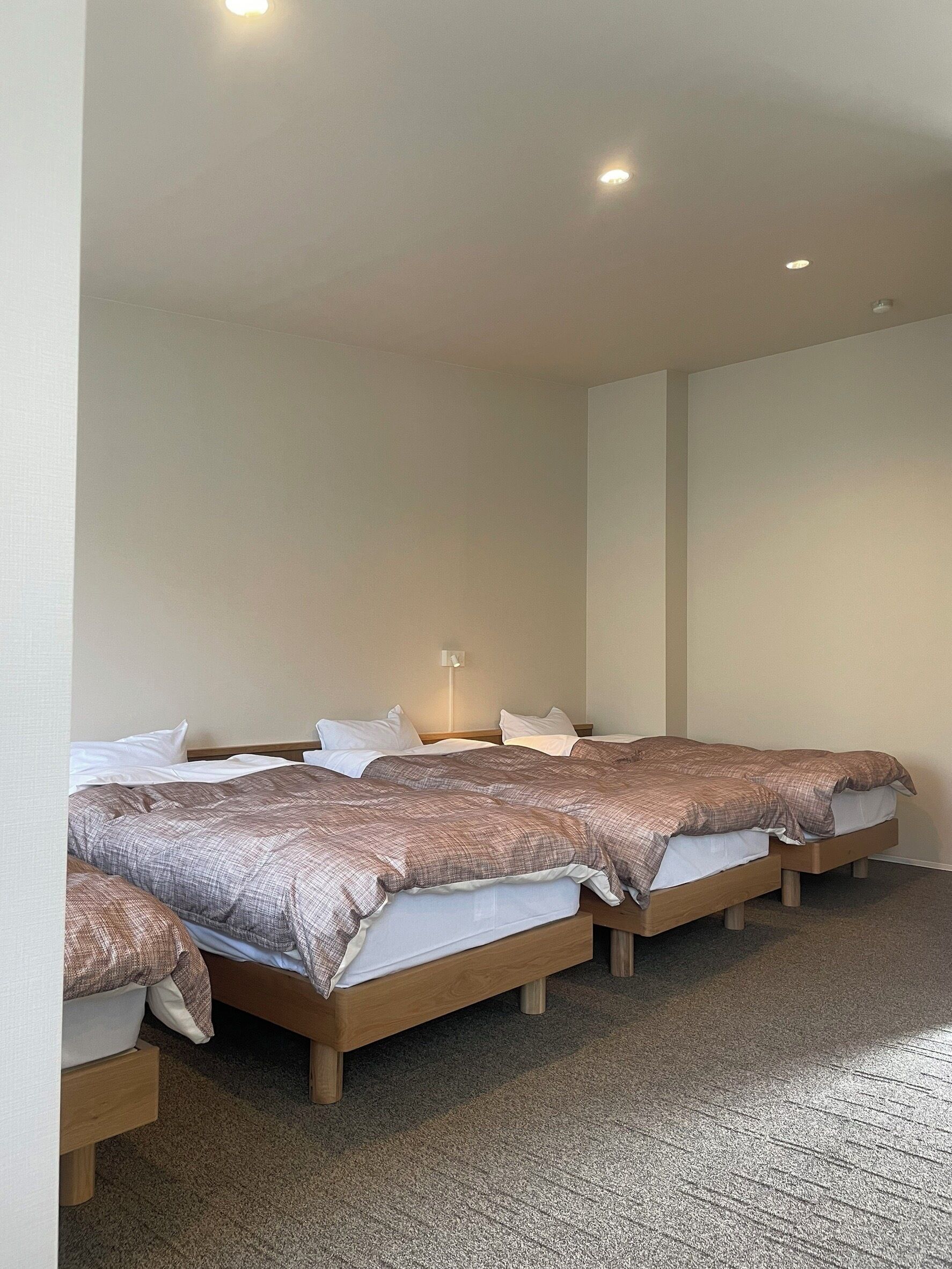 Ein Hotelzimmer mit vier Einzelbetten, einem Nachttisch, einer an der Wand befestigten Lampe und einem Teppichboden.