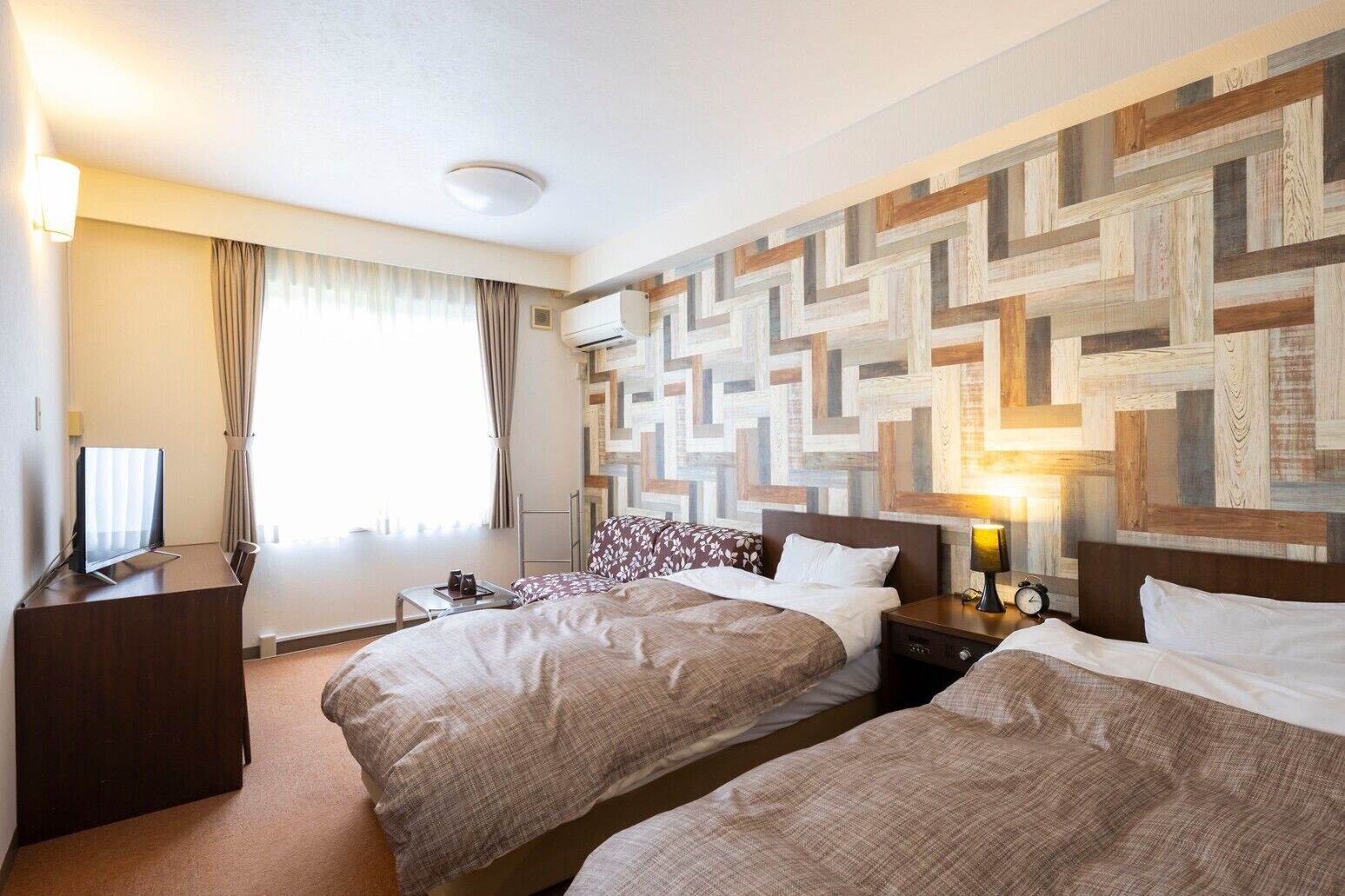 Une chambre d’hôtel avec deux lits, un bureau, une télévision, une fenêtre avec des rideaux et un mur orné d’un motif géométrique.