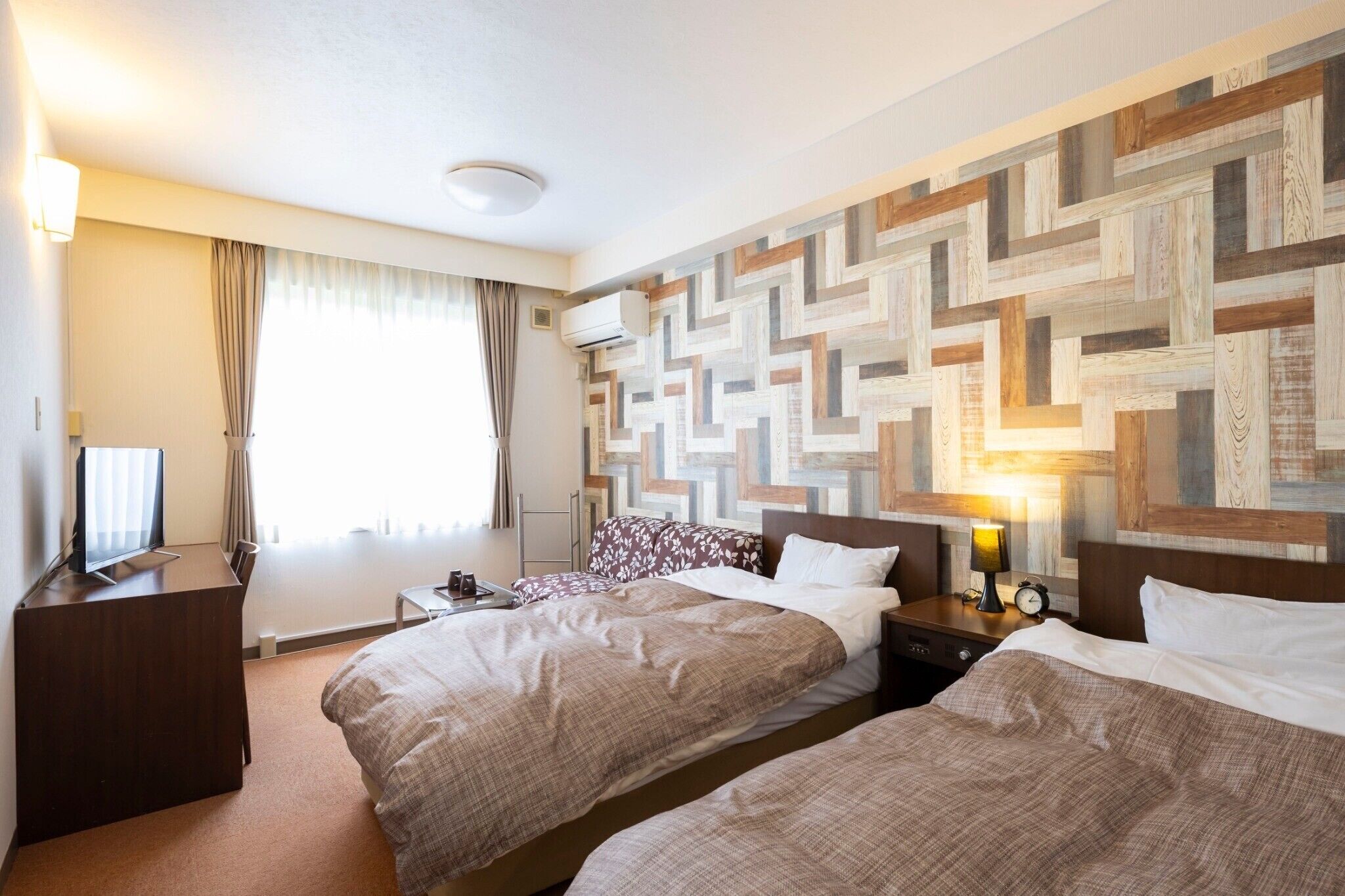 Ein Hotelzimmer mit zwei Betten, einem Fernseher, einem Schreibtisch, einer Lampe und einer Wand mit geometrischem Muster.