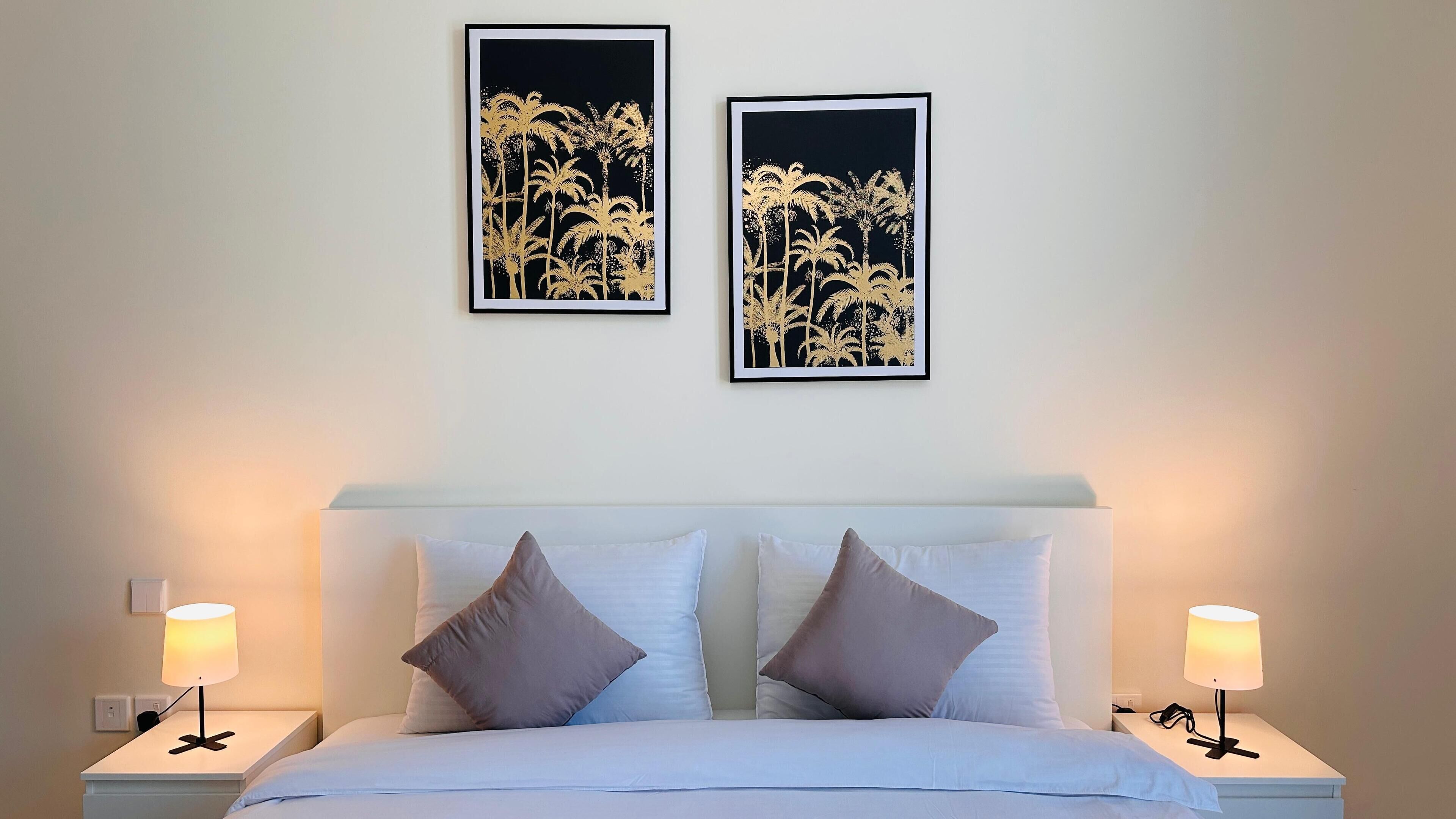 A neatly made bed with white bedding and two pillows, flanked by nightstands with lamps. Two framed artworks depicting palm trees are hung above the bed.