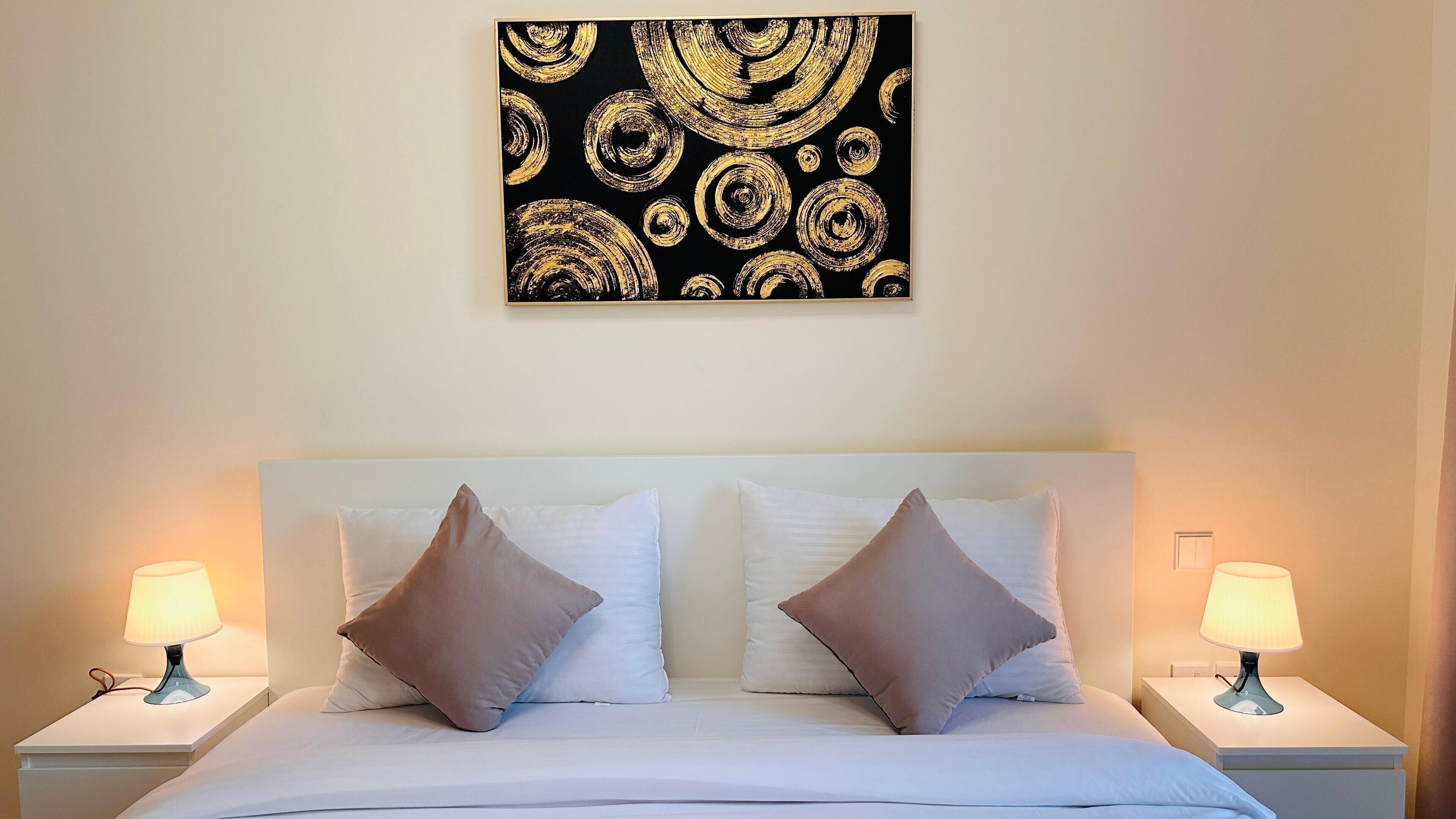 Habitación de hotel con cama blanca, dos mesitas de noche, dos lámparas y un cuadro abstracto en tonos negros y dorados en la pared.