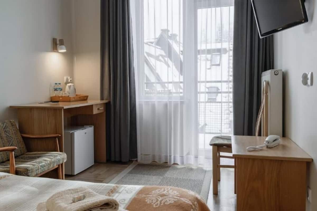 A hotel room with a bed, a desk, a chair, a small refrigerator, a telephone, and a TV mounted on the wall.