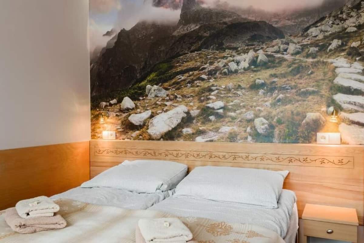 A bedroom with a mountain view mural, a wooden headboard, two beds with white linens, and bedside lamps.