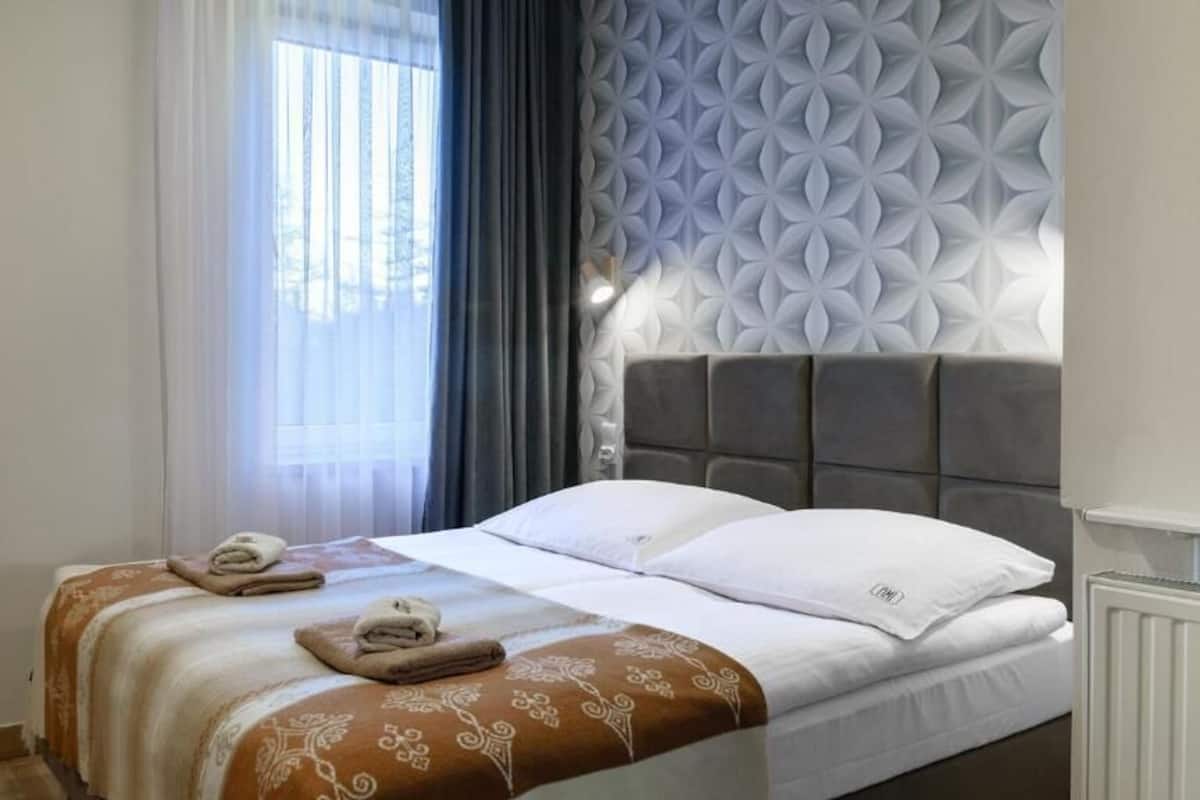 A neatly made bed with a patterned bedspread, a headboard with a geometric design, and a window with sheer curtains.