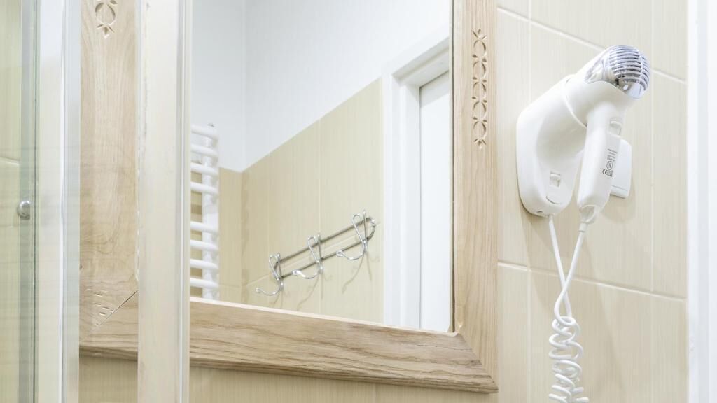 Casa de banho com secador de cabelo fixado na parede, porta-toalhas e espelho com moldura de madeira.