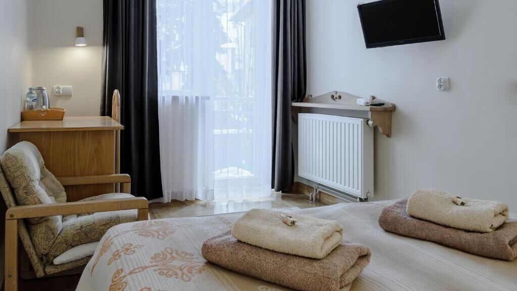 Habitación de hotel con dos camas, una silla de madera, un escritorio con lámpara, un televisor y una ventana con cortinas.
