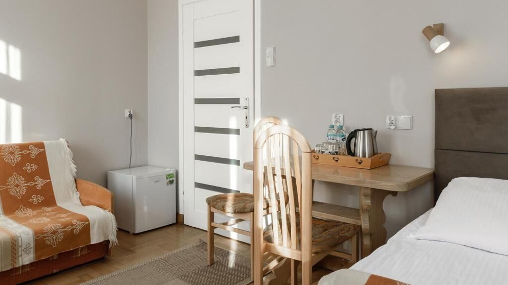 A compact hotel room with a bed, a sofa, a wooden chair, a small table with a kettle and bottles, and a white door with horizontal stripes.