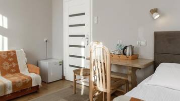 A compact hotel room with a bed, a sofa, a wooden chair, a small table with a kettle and bottles, and a white door with horizontal stripes.