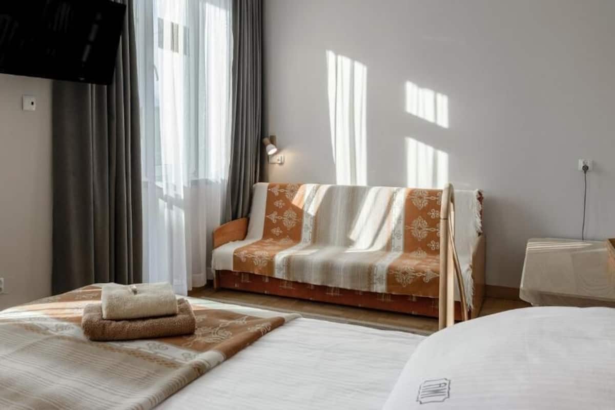 A hotel room with a bed, a sofa, a TV mounted on the wall, and a window with curtains.