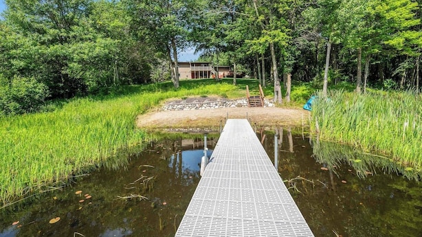 Une jetée en bois menant à une maison entourée d’une végétation luxuriante et bordée d’un étang.
