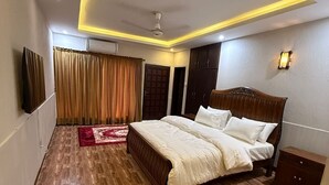 A bedroom with a wooden floor, a bed with white bedding, a ceiling fan, and a TV mounted on the wall. - Sundas Lodge (Islamabad)