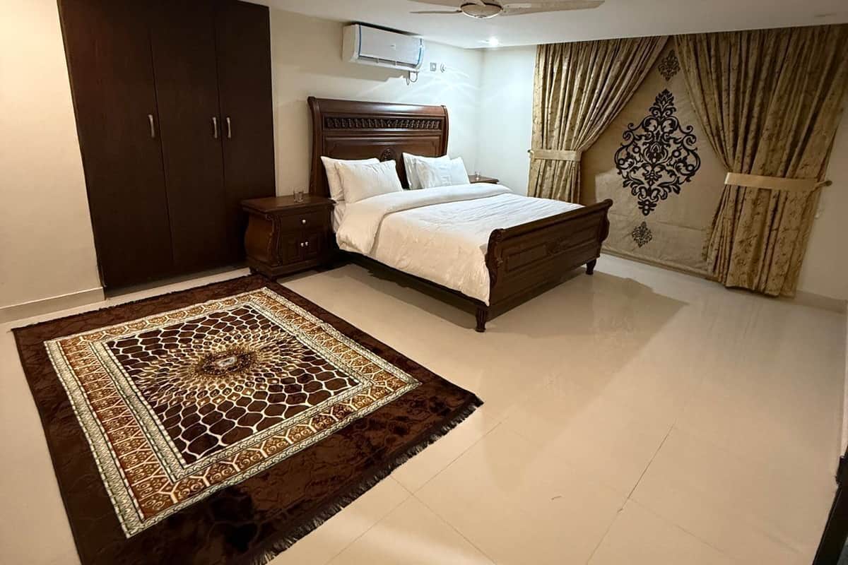 A bedroom with a bed, a wardrobe, a ceiling fan, an air conditioning unit, and a patterned rug.
