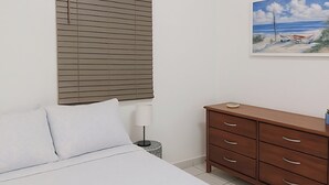 Une chambre à coucher avec un lit, une commode en bois, un tableau représentant une scène de plage et une fenêtre avec des stores.
