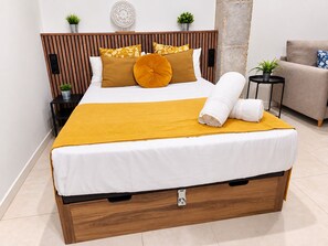 A neatly made bed with a white mattress, a mustard-colored throw blanket, and a rolled white towel.