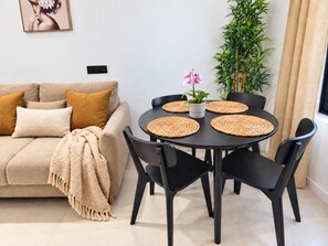 A living room with a beige sofa, black chairs, a round black table with woven placemats, a potted plant, and a framed picture on the wall.