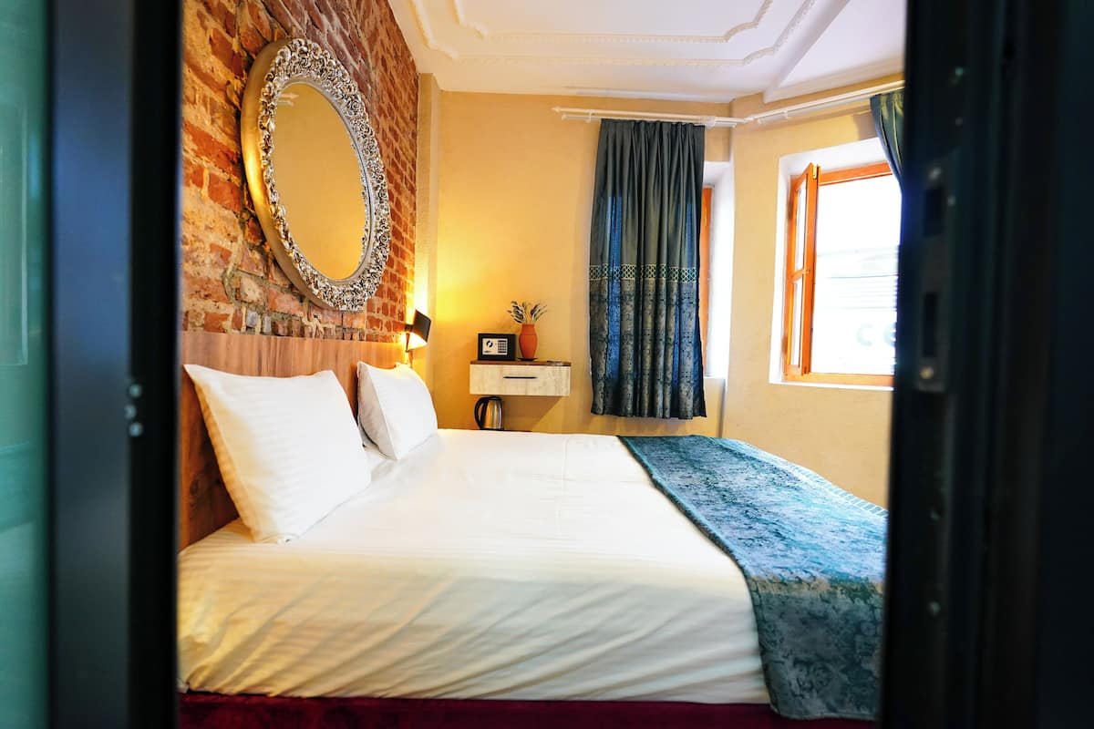 A hotel room with a brick wall, a large mirror, a bed with white linens, a nightstand with a lamp, and a window with curtains.