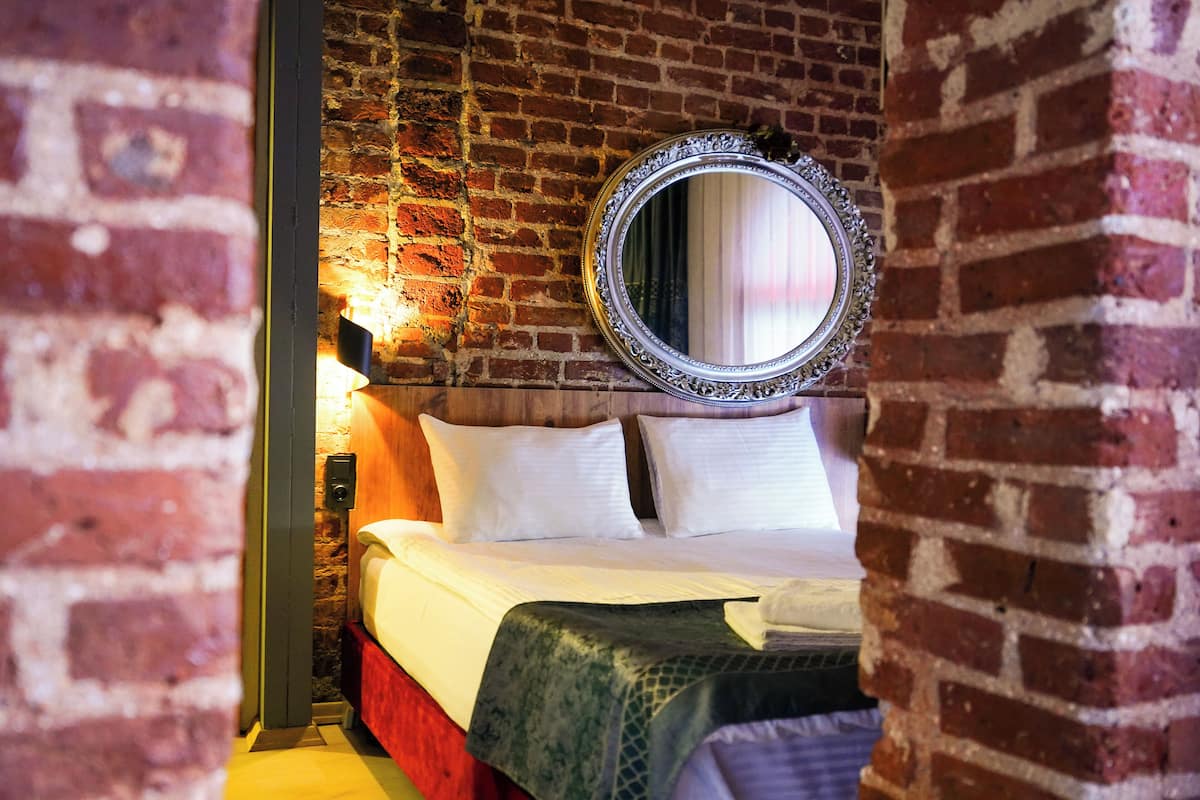 A room with a brick wall, a bed with white bedding, a green throw blanket, and a mirror with an ornate frame.