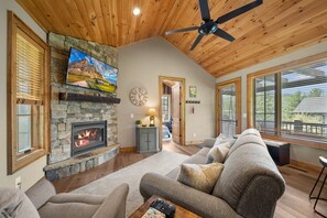 A cozy living room with a stone fireplace, a ceiling fan, and large windows. - River Rock - 1mile to downtown Banner Elk, pets (Banner Elk)