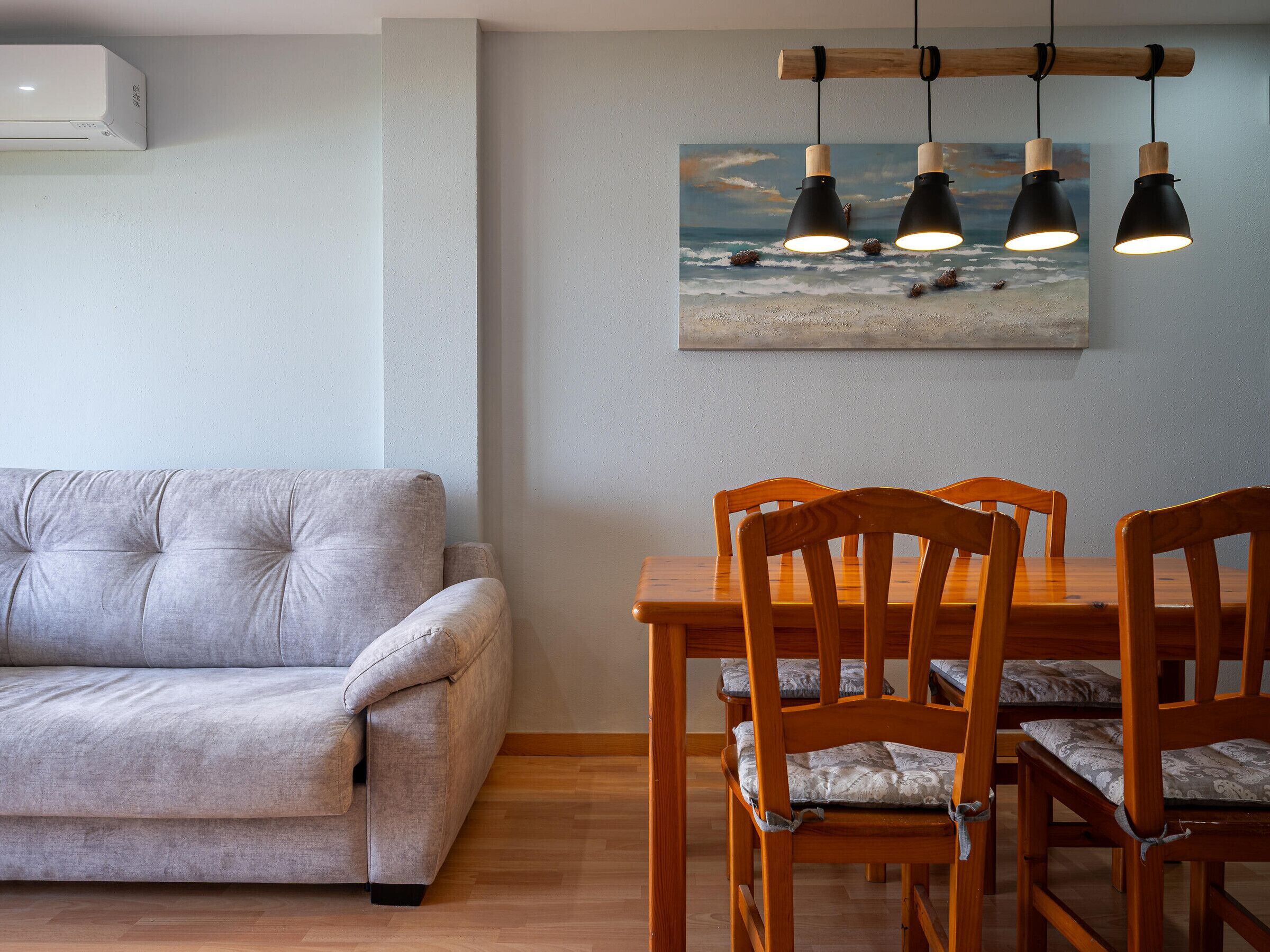 Ein Wohnzimmer mit grauem Sofa, einem Holzes Esstisch und Stühlen. An der Wand hängt ein Bild von einer Strandansicht.