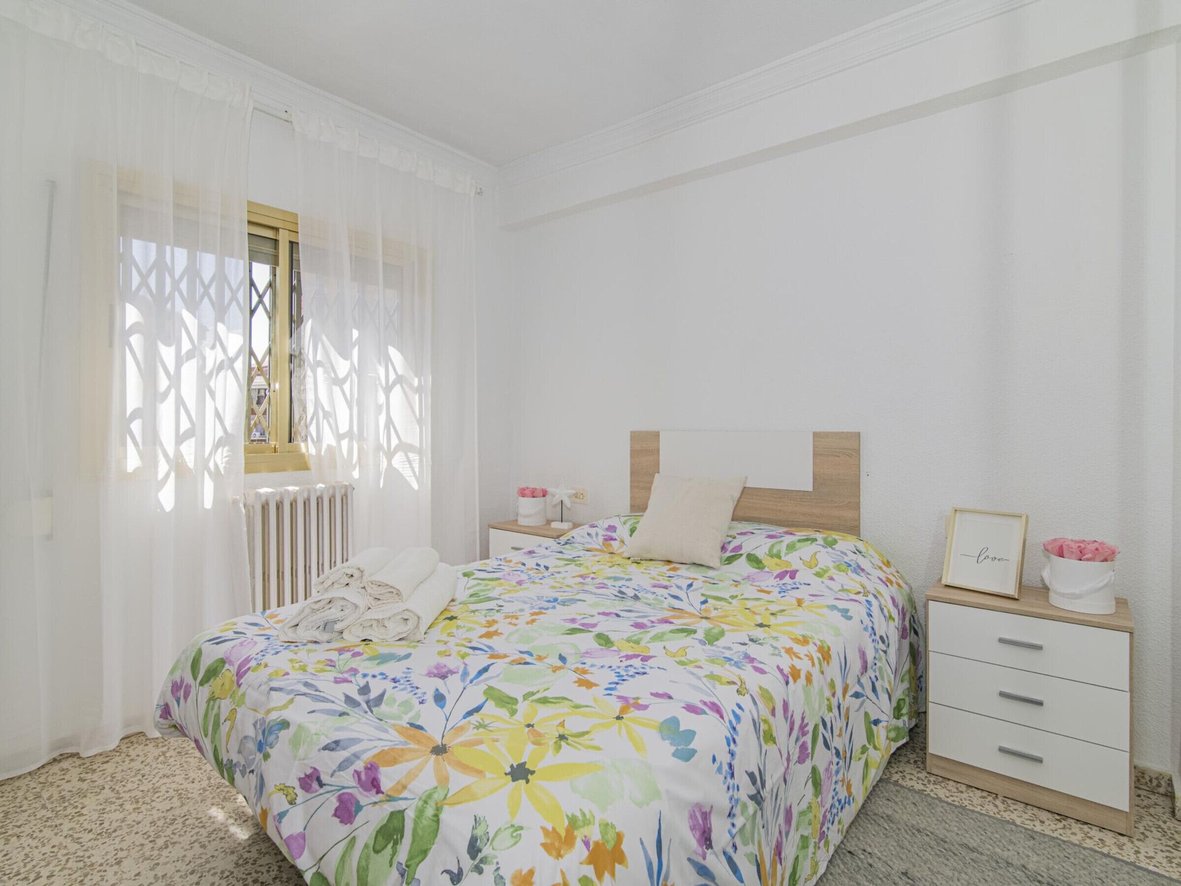 A bedroom with a floral bedspread, a wooden headboard, a bedside table with a picture frame, and a radiator.