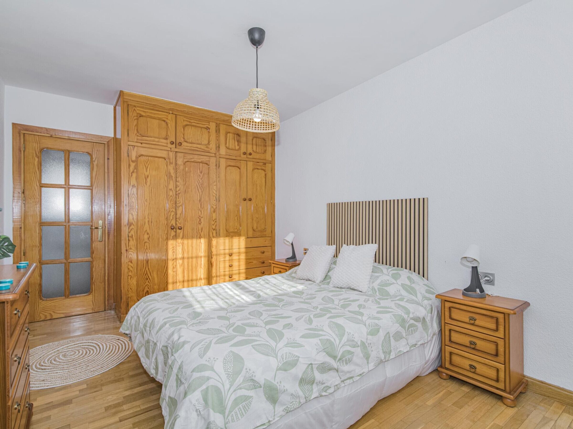 A bedroom with a bed, wooden wardrobe, bedside table, lamp, and a hanging light fixture.