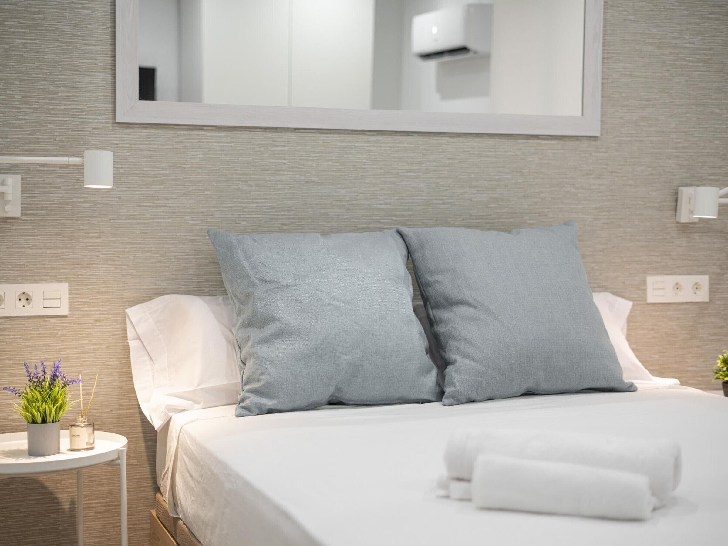 Camera d'hotel moderna con un letto rifatto, un piccolo tavolino con una pianta e uno specchio sopra il letto.