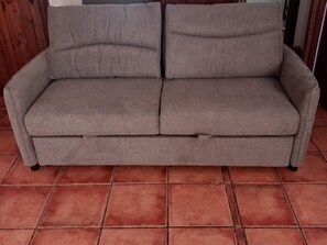A grey sofa with a cushion on a tiled floor.