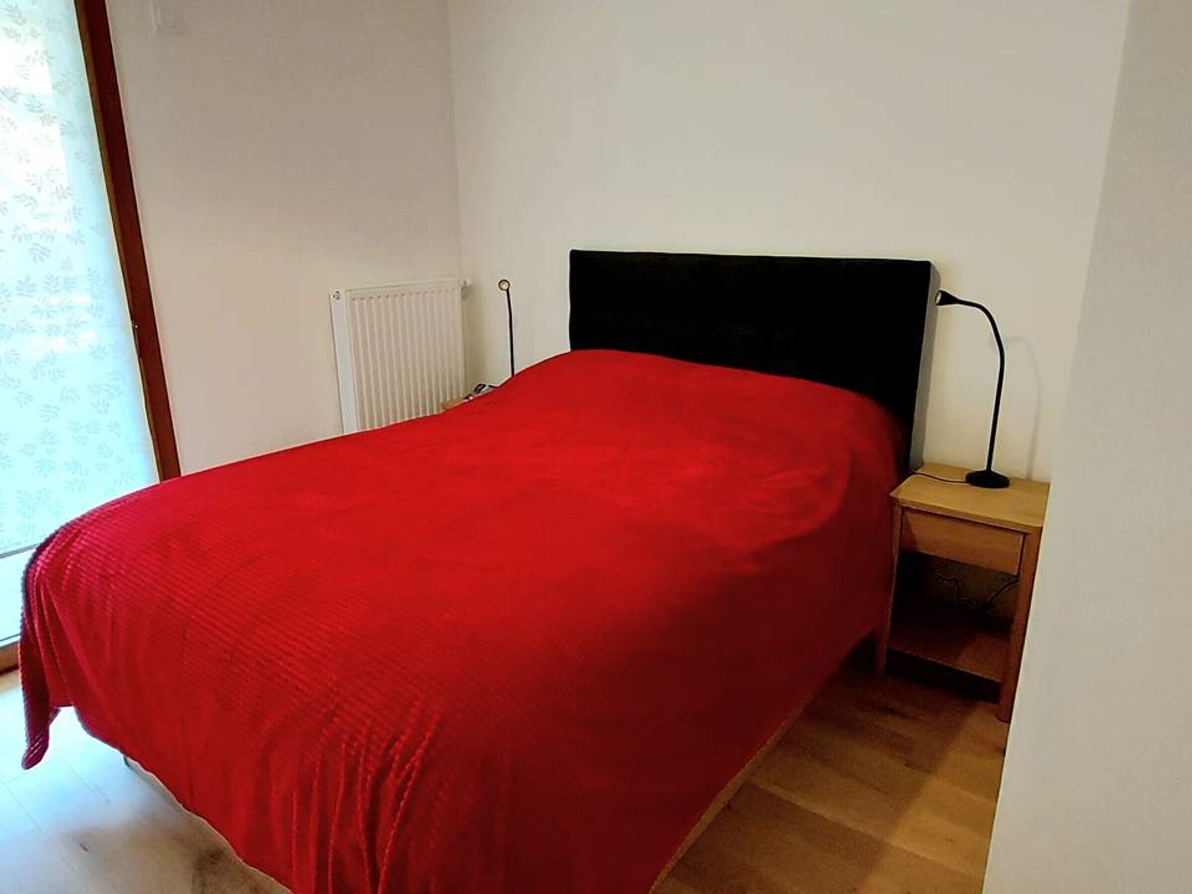 A single bed with a red bedspread in a room with white walls, a radiator, and a nightstand with a lamp.