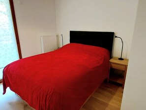 Un llit individual amb un estovat vermell en una habitació de parets blanques, un radiador i una nitu amb una làmpada.