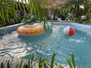 A swimming pool with inflatable rings and a ball, surrounded by palm trees and a tiled edge.