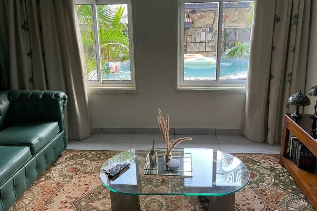 A living room with a glass coffee table, a sofa, and two windows with views of a pool and outdoor area.