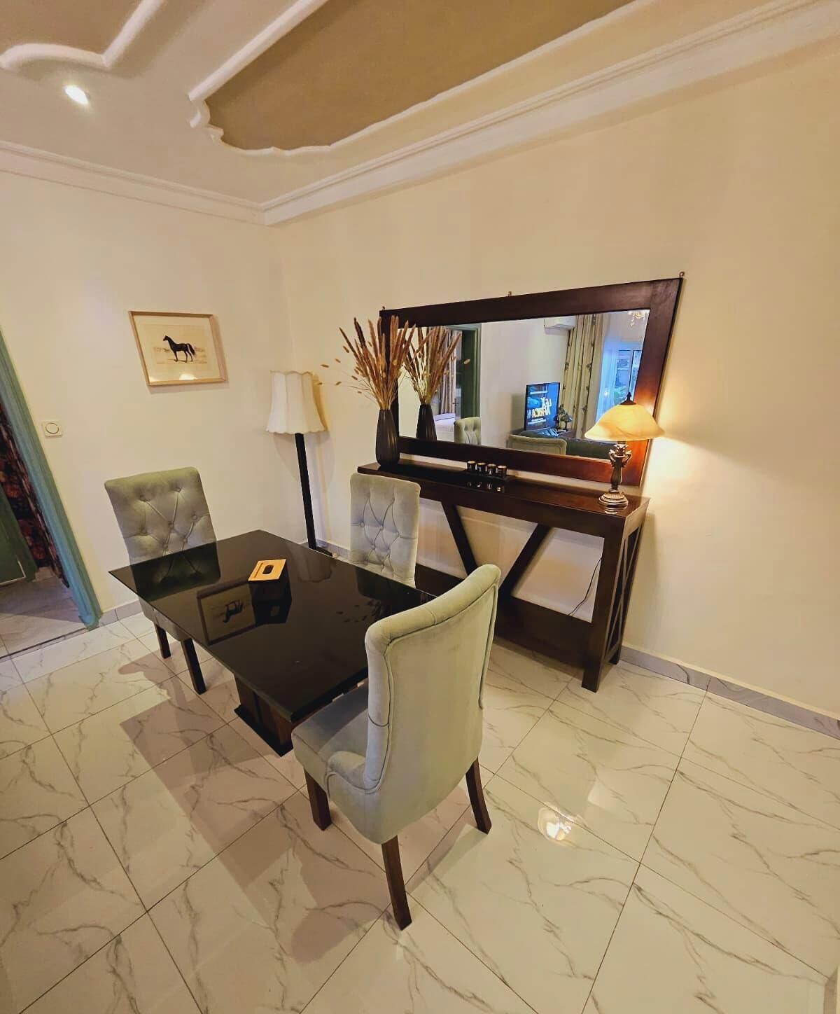 A dining area with a glass-top table, two upholstered chairs, a dark wooden sideboard, a mirror, and a lamp.