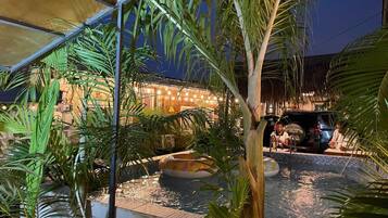 Un área de piscina interior con palmeras, una silla de descanso y una estructura con techo de paja.