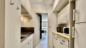 A narrow kitchen with white cabinets, a stainless steel range hood, a microwave, and a refrigerator.