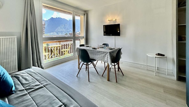 A modern hotel room with a large window offering a mountain view, a dining area with a table and chairs, a flat-screen TV mounted on the wall, and a cozy bed with a blue pillow.