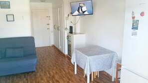 Une pièce avec un canapé bleu, une table recouverte d’une nappe à motifs, un téléviseur fixé au mur et un calendrier accroché au mur.