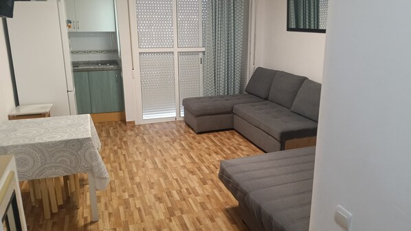 A living room with a grey sectional sofa, a small table with a patterned tablecloth, and a kitchen area with cabinets and a window with patterned curtains. - Studio '100m from the Beach' with Shared Pool, Private Terrace (Matalascañas)