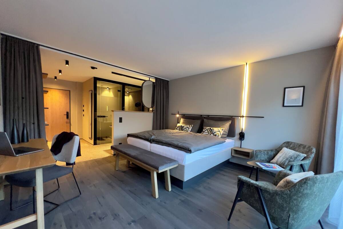 A modern hotel room with a large bed, a desk, a chair, and a bedside table.