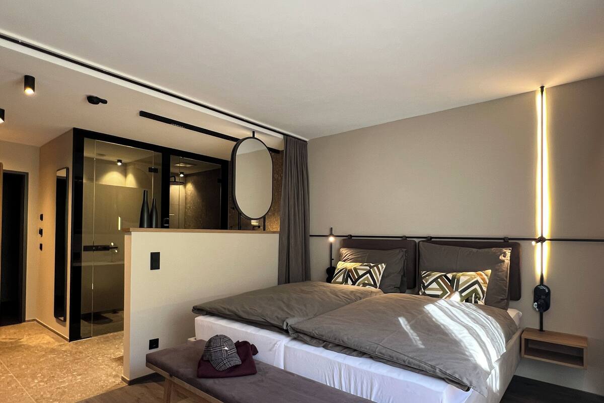 A modern hotel room with a large bed, a bench, a mirror, and a bedside table.