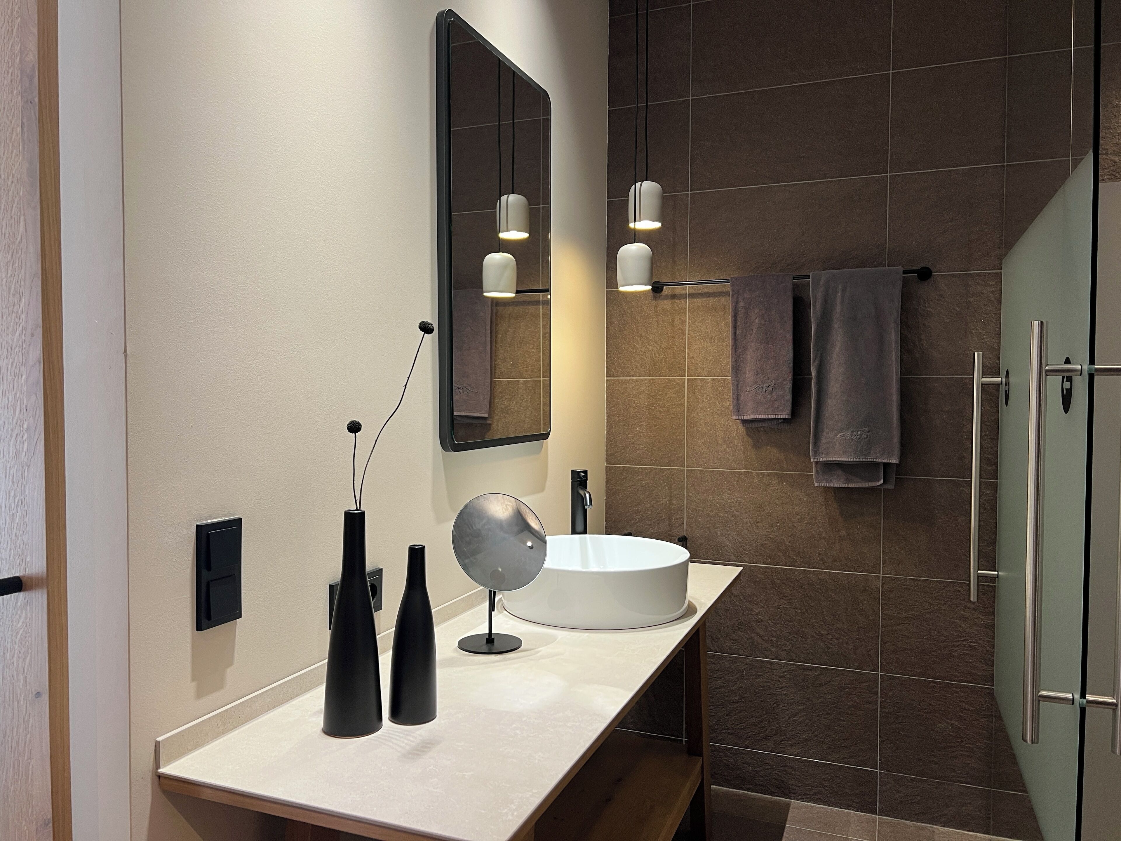A modern bathroom with a white sink, two black vases, a round mirror, and a wall-mounted light fixture.