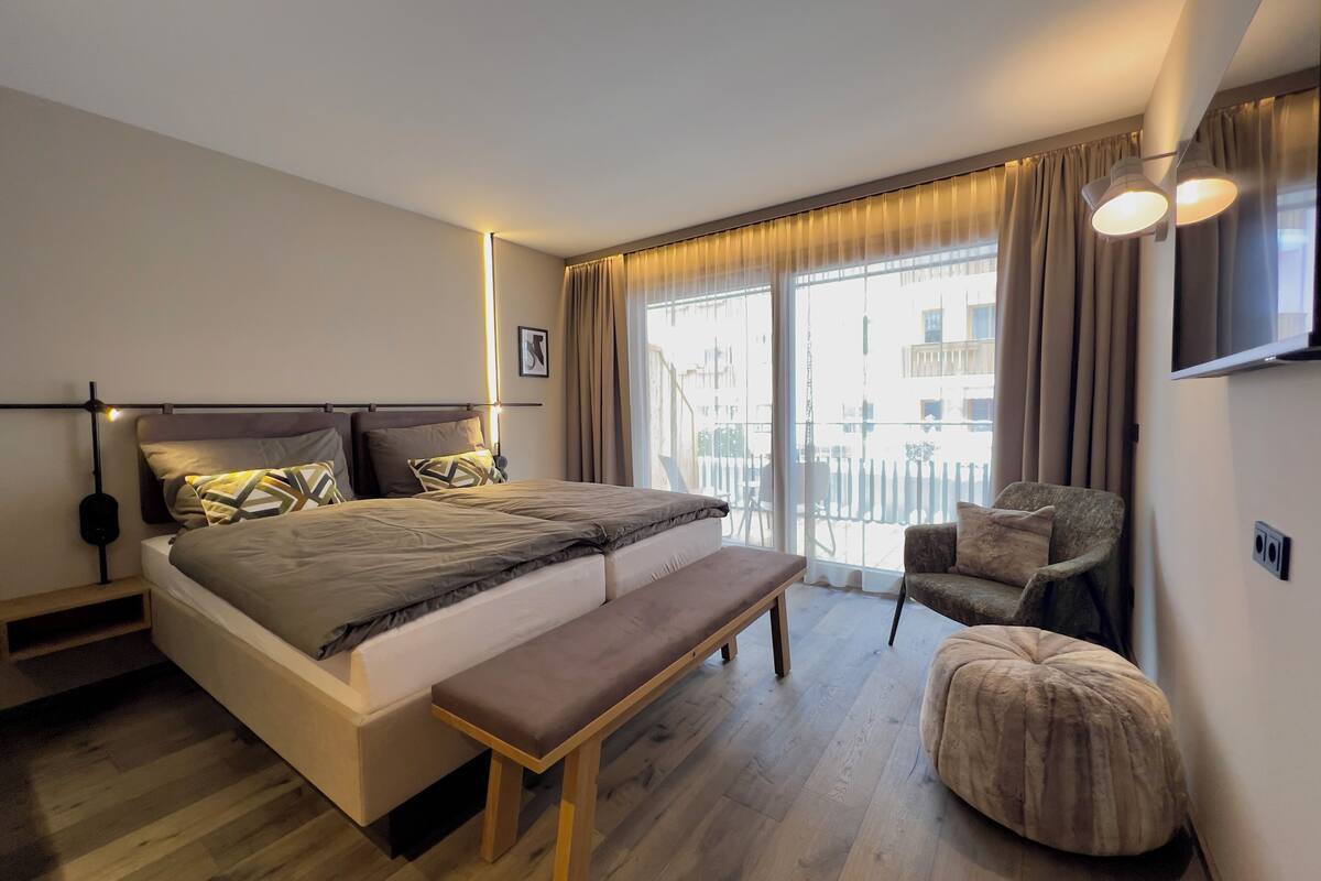 A modern bedroom with a large bed, bedside tables, a bench, a chair, and a TV mounted on the wall.
