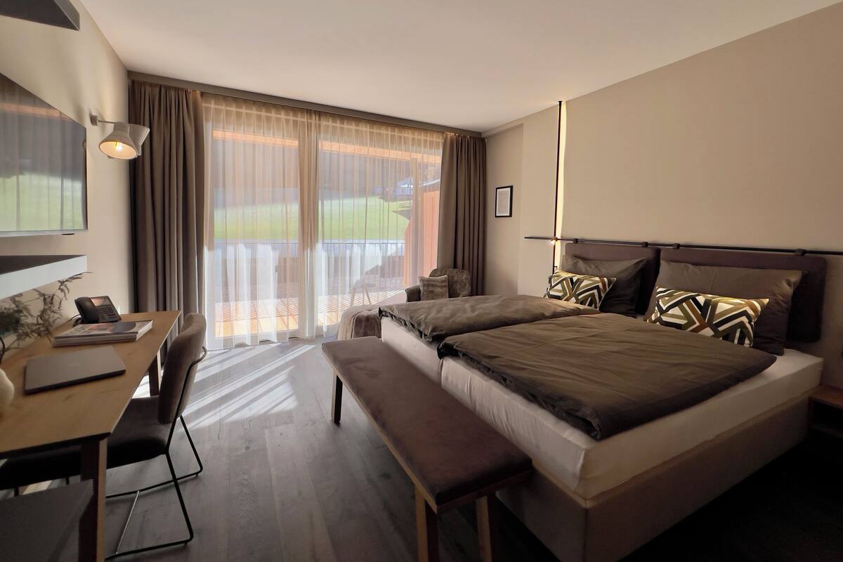 A hotel room with a large bed, a desk with a chair, a bench, and a view of a green landscape through the window.