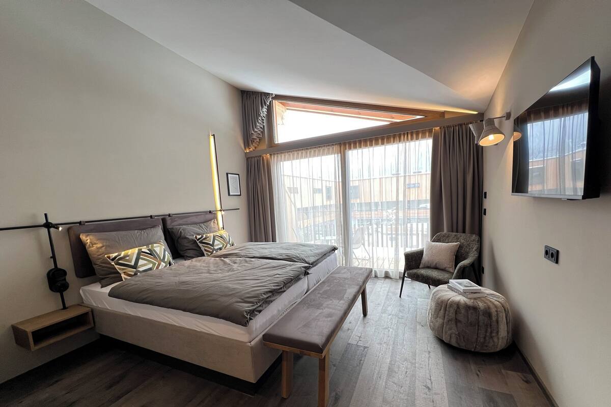 A modern bedroom with a large bed, a bench, a chair, and a TV mounted on the wall.